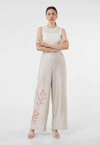 Jasnobeżowe spodnie o szerokich nogawkach z czerwonym haftem w liście po lewej stronie, w połączeniu z białym bezrękawnikowym topem z prążkowanej tkaniny.
