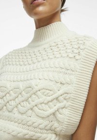 Crème kleurige gebreide trui met een hoge ribbelkraag en een textuur van kabelpatronen over de voorkant. Mouwloos ontwerp met een zachte afwerking.