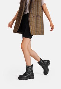 Stivaletti neri in pelle con suola spessa, design con lacci e cinturini decorativi sulla parte superiore. Indossati con un gilet a quadri e pantaloni corti neri.