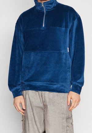 Man draagt een blauw fleece-trui met lange mouwen en een halve ritssluiting en lichtgrijze cargobroek, staand tegen een effen achtergrond.