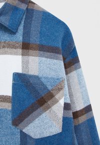 Chemise en laine à carreaux bleue, blanche et marron avec une poche poitrine et un col plié sur un fond clair.