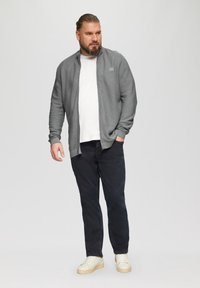 Graue Zip-Jacke mit strukturiertem Stoff über einem weißen T-Shirt, kombiniert mit dunklen Jeans und weißen Turnschuhen. Mann steht lässig.