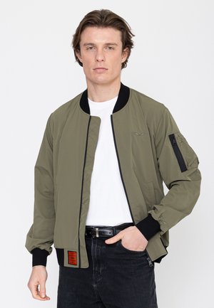 Jeune homme portant une veste bomber vert olive avec des bordures noires, un t-shirt blanc, un jean noir, debout devant un fond blanc uni.