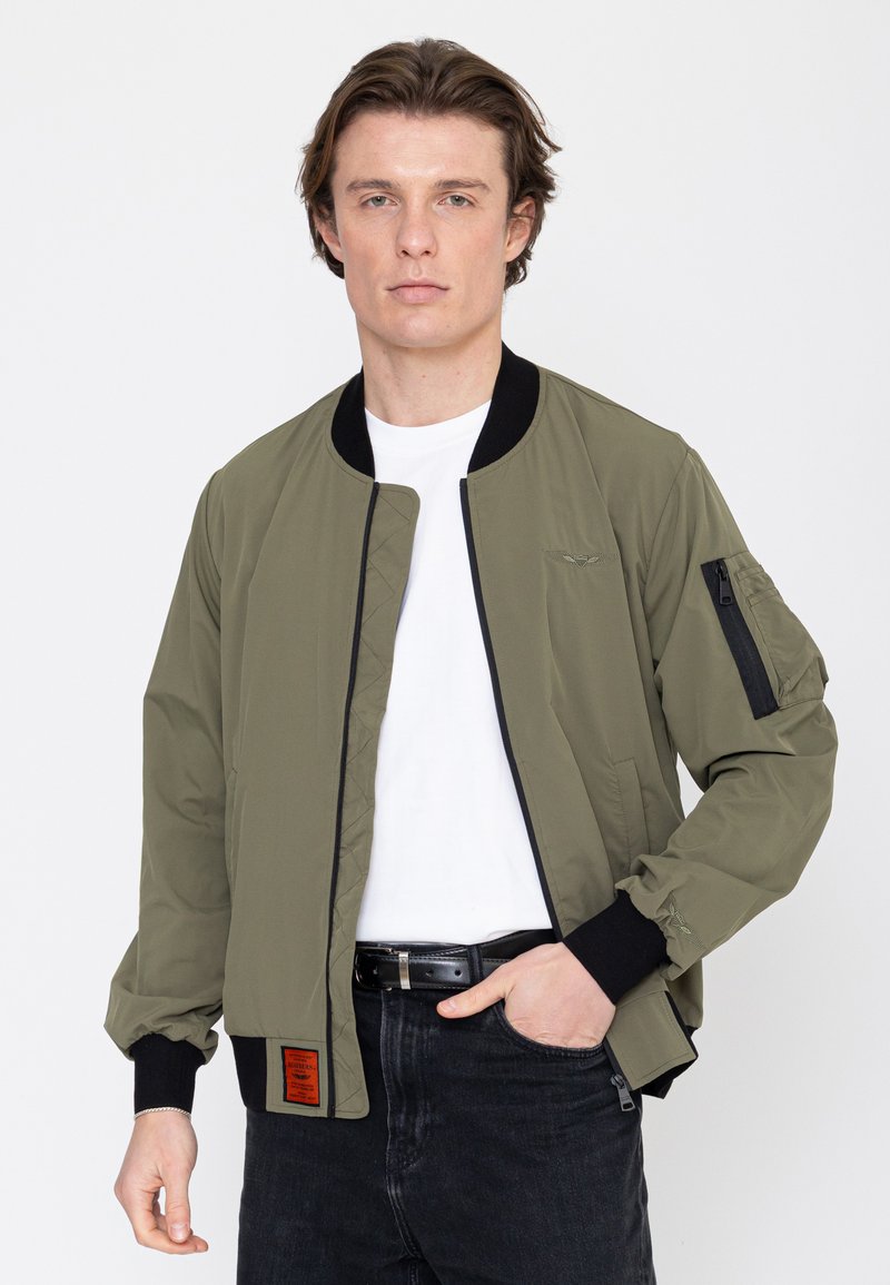 Junger Mann, der eine olivgrüne Bomberjacke mit schwarzem Rand, ein weißes T-Shirt und schwarze Jeans trägt, vor einem schlichten weißen Hintergrund steht.