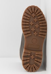 Semelle d'une botte Timberland avec un épais motif de bande de roulement marron comportant des croix et le logo de la marque, inclinée sur un fond blanc à coins.