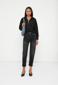 Felpa nera con zip, jeans scuri e tacchi neri. Una borsa blu è tenuta in mano, con una cintura aderente e uno stile casual. Tessuto dalla consistenza morbida.