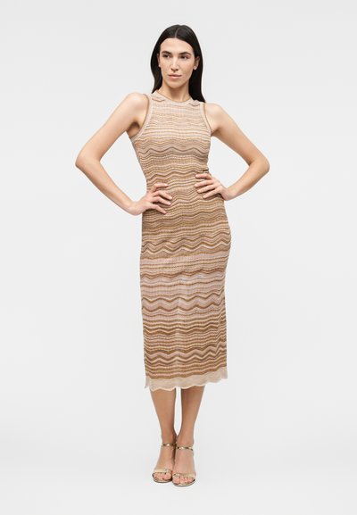 Γυναίκα με αμάνικο, μέχρι το γόνατο φόρεμα με κυματιστό μοτίβο σε μπεζ και καφέ αποχρώσεις, φορώντας χρυσά πέδιλα με λουράκι στον αστράγαλο, στέκεται με τα χέρια στη μέση.