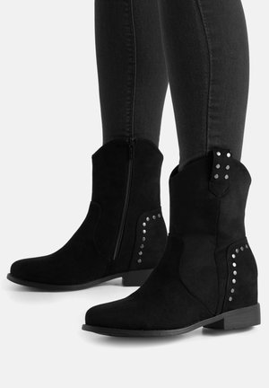 Botas de ante negras hasta la mitad de la pantorrilla con cremalleras laterales y detalles de tachuelas plateadas, usadas con pantalones negros ajustados sobre un fondo neutro.