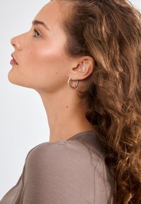 Brincos em argola torcida de prata, usados por uma pessoa com cabelo ondulado, exibindo um tom de pele natural e uma camisa castanha clara com textura canelada.