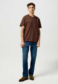 Ruskea puuvillapaita lyhyillä hihoilla, pieni logo rinnassa, yhdistettynä sinisiin farkkuihin ja ruskeisiin saappaisiin.