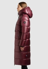 Femme portant un long manteau rembourré bourgogne brillant avec capuche, poches latérales et écussons noirs sur la manche et l'ourlet inférieur, faisant face à la gauche.