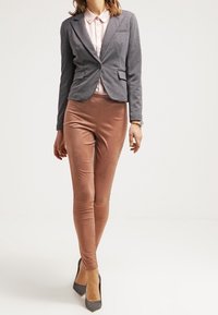 Blazer en tissu gris sur une chemise rose pâle à col, associé à des leggings marron clair brillants et des chaussures à talons gris.