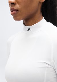 Λευκό τοπ με μακρύ μανίκι και ψηλό γιακά, με ομαλή, ελαστική υφή. Διαθέτει μαύρη λογότυπη λεπτομέρεια στον γιακά και κομψή εφαρμογή.