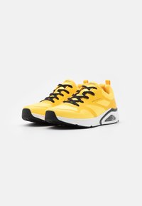 Yellow athletic sneakers with black accents, textured mesh upper, round toe, lace-up design, and a visible air cushioning unit in the sole.