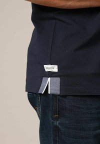 Chemise bleu marine avec une fente sur le côté, présentant un motif à carreaux et une étiquette en tissu sur l'ourlet ; associée à un jean en denim foncé.