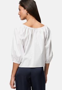 Bílá off-the-shoulder blůza s vypufovanými rukávy, elastickým výstřihem a mírně zkráceným spodním lemem, vyrobená z lehkého bavlněného materiálu.
