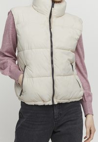 Krämfärgad pufferväst med hög krage, framdragkedja och sidofickor. Har ett quiltat mönster och elastisk nederkant. Bärs över en rosa tröja.