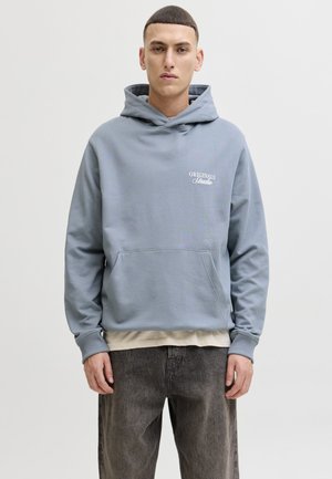 Jonge man met een lichtblauwe hoodie met de tekst "ORIGINALS Studio", een beige overhemd eronder en donkergrijze broek, staand tegen een effen achtergrond.