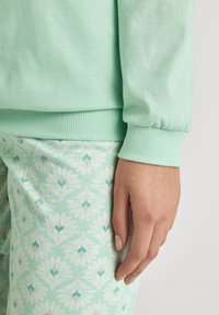 Hellgrüner Sweatshirt mit gerippten Bündchen, kombiniert mit gemusterten mintfarbenen Schlafanzughosen, die weiße Blumenmuster und subtile rosa Akzente aufweisen.