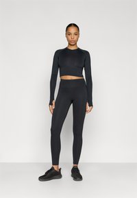 Black long-sleeve cropped top paired with high-waisted black leggings. Textured fabric, fitted design. Model wears black athletic shoes.
