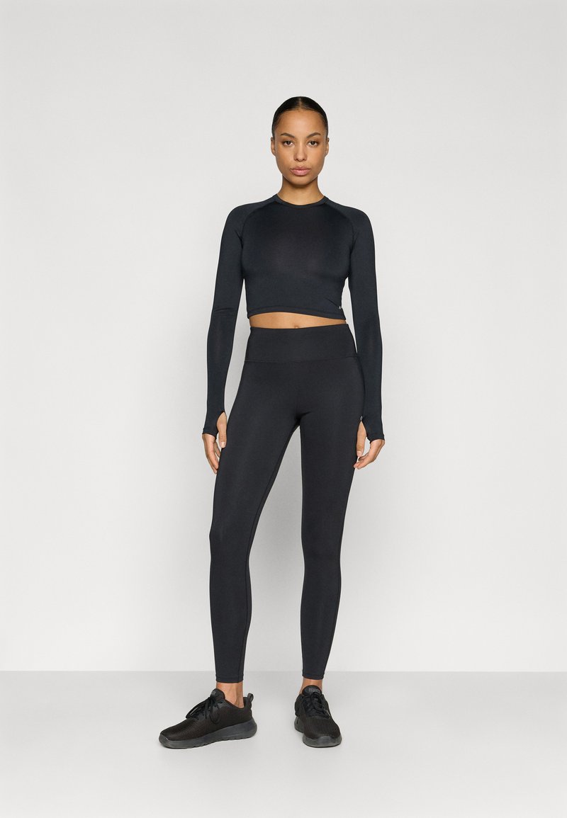 Haut court noir à manches longues associé à des leggings noirs taille haute. Tissu texturé, coupe ajustée. Le mannequin porte des chaussures de sport noires.