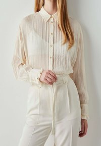 Blouse légère plissée de couleur blanc cassé avec un col classique et une fermeture à boutons, associée à un pantalon blanc taille haute avec des poignets à volants.