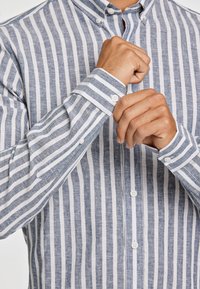 Camicia a righe blu e bianche realizzata in un tessuto leggero. Presenta un colletto con bottoni, maniche lunghe e bottoni visibili lungo la parte anteriore.