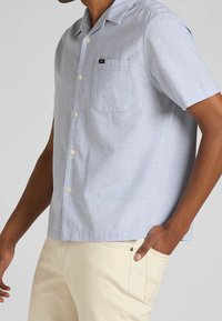 Camicia azzurra a maniche corte con bottoni in un tessuto testurizzato, dotata di tasca sul petto a sinistra e una discreta etichettatura.