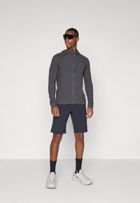 Veste de sport grise zippée avec capuche, tissu texturé et accents réfléchissants, associée à un short noir et des chaussures de sport grises.