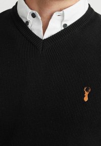 Pull noir côtelé à col en V superposé sur une chemise blanche à boutons. Présente un petit logo de cerf orange sur le côté gauche de la poitrine.