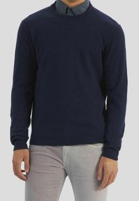 Donkerblauwe crewneck sweater gemaakt van zachte gebreide stof. Bevat lange mouwen en een aansluitend ontwerp, gedragen over een gestreept overhemd met kraag.