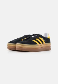 Zapatillas de ante negras con rayas amarillas, detalles en blanco y una suela de goma. Presentan un diseño de cordones planos y soporte acolchado en el tobillo.