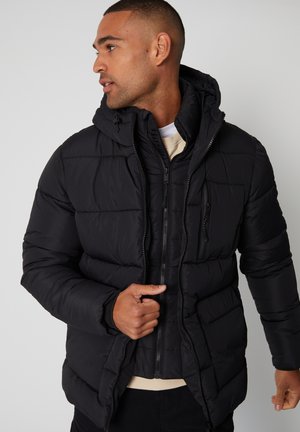 Schwarze Pufferjacke mit Kapuze, ausgestattet mit einem gesteppten Design, Reißverschluss und seitlichen Taschen. Das Material scheint weich und isoliert zu sein.