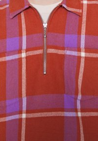 Gros plan sur une chemise à carreaux rouge et violette avec une fermeture éclair argentée à moitié fermée, dotée d'un col et d'une poche poitrine, portée par-dessus un sous-vêtement blanc.