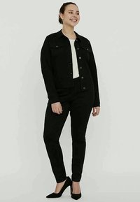 Zwarte denimjas met knoopsluiting en twee borstzakken, gecombineerd met zwarte broek en puntige zwarte hakken, tegen een lichte achtergrond.