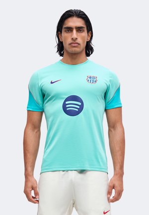 Hombre con cabello oscuro hasta los hombros, vistiendo una camiseta turquesa del FC Barcelona y pantalones cortos blancos sobre un fondo liso.