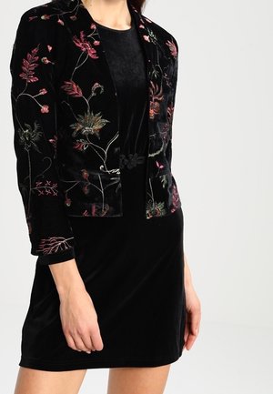 Femme portant une robe en velours noir avec une veste courte noire ornée de broderies florales roses et vertes.