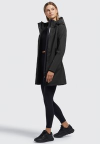 Veste de pluie noire imperméable avec capuche, dotée de boutons pression, de poches zippées et d'une coupe décontractée, associée à un legging noir et à des baskets.