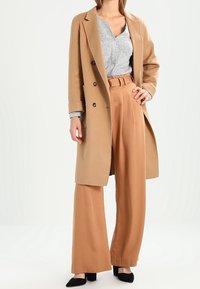 Abrigo entallado de color marrón claro con grandes botones, llevado sobre un suéter gris y pantalones beige de pierna ancha a juego. Unos zapatos planos negros de punta completan el conjunto.
