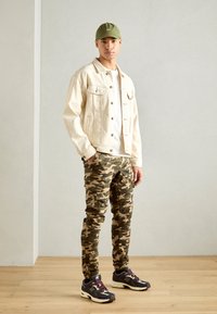 Beżowa dżinsowa kurtka na białym T-shircie, zestawiona z kamuflażowymi spodniami cargo i czarnymi trampkami z fioletowymi akcentami. Model stoi na jasnym tle.