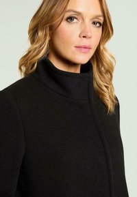 Abrigo negro de lana con cuello alto, corte recto y diseño minimalista. Presenta una superficie texturizada y detalles de costuras limpias y ocultas.