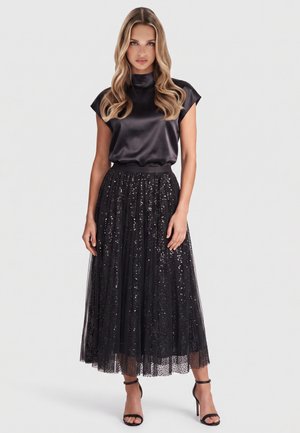 Woman with long wavy hair wearing a short-sleeve black satin top and a black sequined tulle midi skirt with black ankle-strap heels.