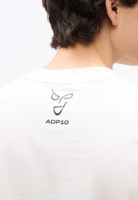 Άτομο που φοράει λευκό πουκάμισο με μαύρο αφηρημένο λογότυπο προσώπου και το κείμενο "ADP10" στο πάνω μέρος της πλάτης κοντά στον λαιμό.