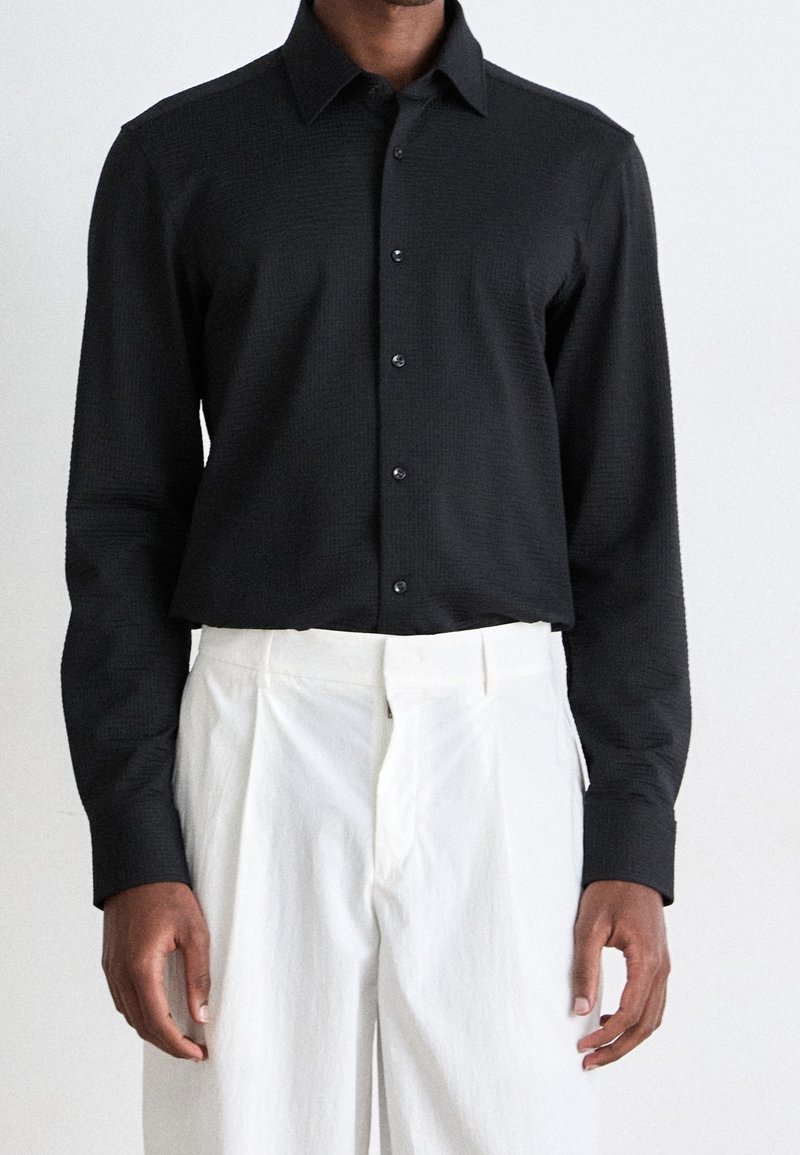 Homme portant une chemise noire à manches longues boutonnée rentrée dans un pantalon blanc plissé taille haute, sur un fond uni.