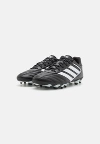 Botas de fútbol negras hechas de cuero sintético, con tres franjas blancas en el lateral, parte superior texturizada y tacos de goma moldeados.