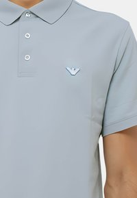 Sivá polo košeľa s klasickým golierom, trojica gombíkov a malým modrým vyšívaným logom na ľavej strane hrudníka. Hladká textúra látky.