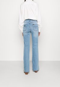 Person, der hellblaue gerade geschnittene Jeans und ein weißes Langarmhemd trägt, von hinten vor einem schlichten weißen Hintergrund betrachtet.