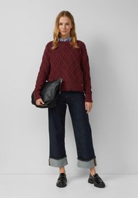 Burgundowy sweter dzianinowy z diamentowym wzorem, luźny krój, ściągacze przy mankietach, zestawiony z szerokimi ciemnymi dżinsami z zagnieceniem na końcu, czarna torebka na ramię, loafersy.