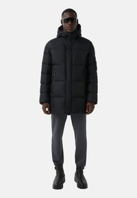 Schwarze Winterjacke mit Kapuze, mit gestepptem Design, Reißverschlusstaschen und geradem Schnitt, kombiniert mit grauen Hosen und schwarzen Stiefeln.