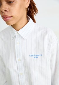 Camisa a rayas blancas con líneas verticales azules, diseño abotonado y logo bordado "Carhartt WIP" en el lado izquierdo. Textura de algodón suave.